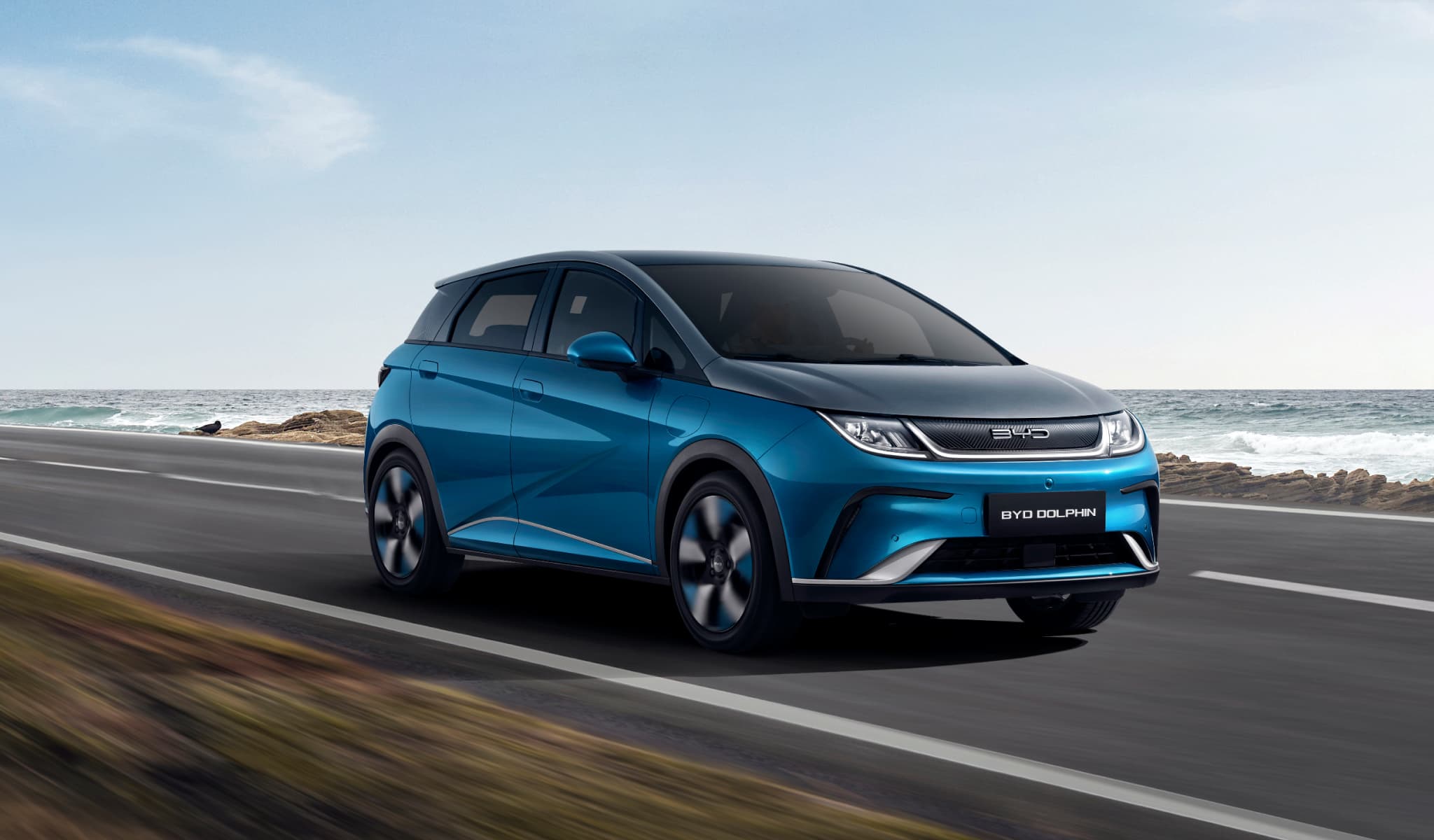 Cotxe elèctric blau BYD Dolphin circulant per una carretera costanera sota un cel clar